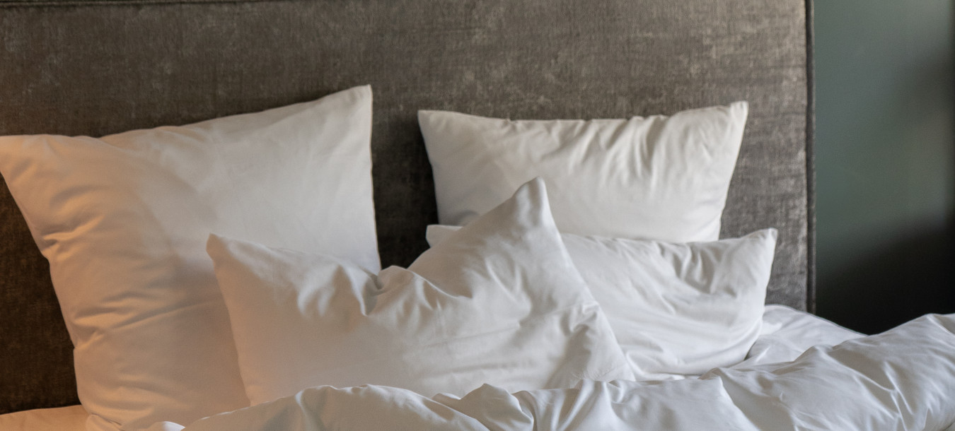 Ein gemütlich-unordentliches Bett mit weißen Kissen und einer weißen Bettdecke vor einer dunkelgrünen Wand.