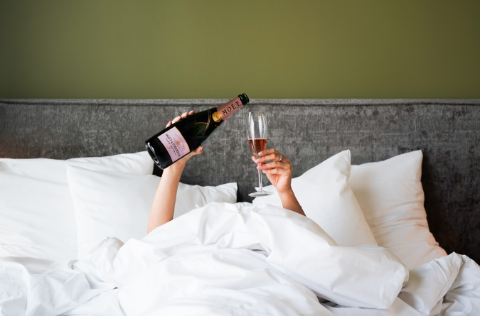 Eine Person, die unter weißen Decken im Bett liegt, hält eine Flasche Champagner und ein mit Champagner gefülltes Sektglas vor einem grauen Kopfteil und einer grünen Wand hoch.