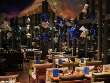 Ein mit blauen und silbernen sternförmigen Ballons dekoriertes Restaurant, das mit Tischen und Glasfenstern für eine Abendveranstaltung gerüstet ist.