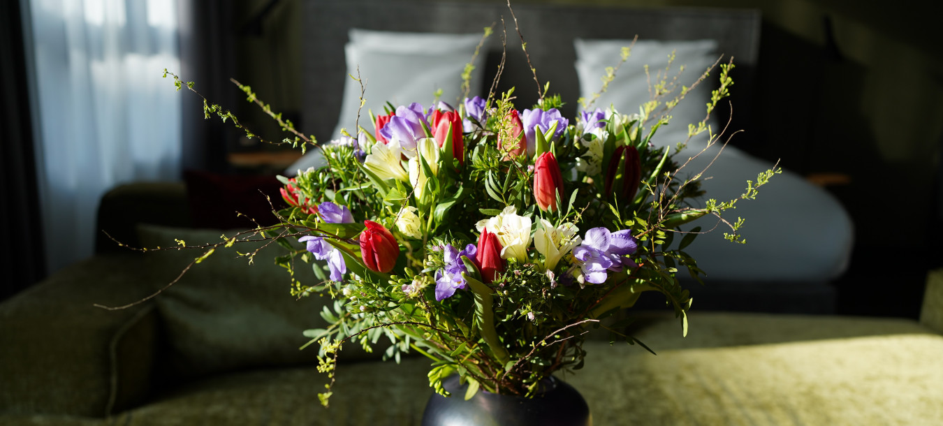 A bouquet of colorful flowers in a dark vase sits on a wooden table in a sunlit room with a bed and green sofa in the background.