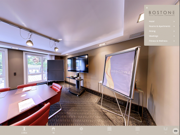 Moderner Besprechungsraum im Boston Hotel mit roten Stühlen um einen Tisch, einem großen Fernseher an der Wand, einem Flipchartständer und großen Fenstern, die Tageslicht hereinlassen. Am Bildrand ist die Navigation des Roomtour zu sehen.
