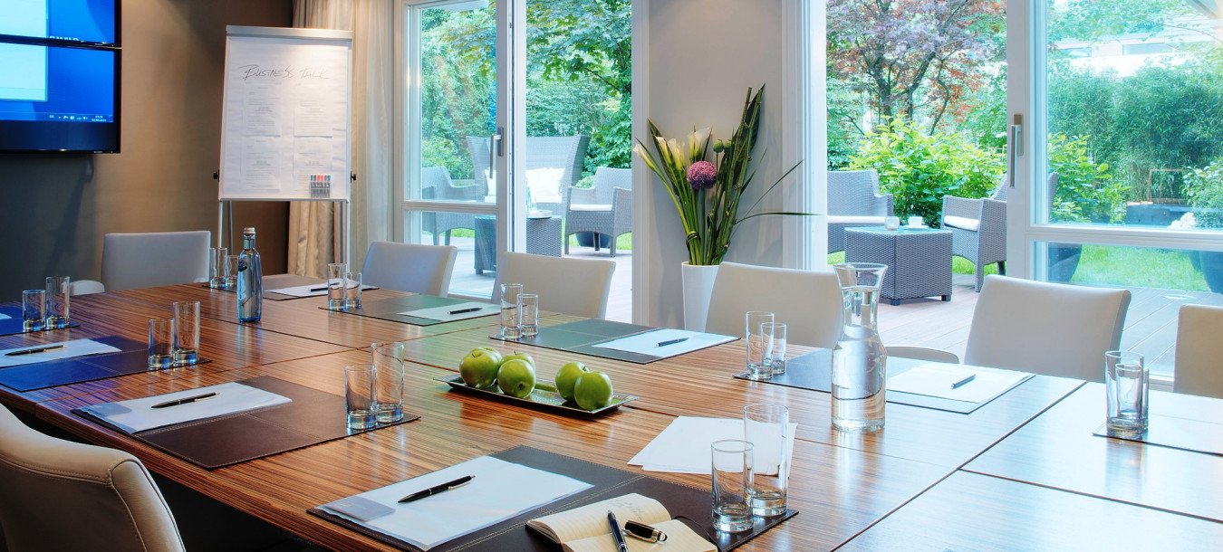 A modern conference room with a large table, notepads, water bottles, and windows overlooking a green garden patio.