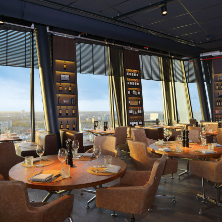 Erleben Sie den modernen Charme des clouds Restaurants mit runden Holztischen und bequemen Lederstühlen unter weitläufigen Panoramafenstern. Die mit Flaschenregalen geschmückten Wände spiegeln eine himmlische Atmosphäre wider – ein Gefühl, a