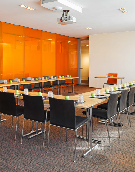 Ein moderner east Konferenzraum mit schwarzen Stühlen um U-förmig angeordnete Tische. Jeder Platz ist mit einem Glas, einem Notizblock, einem Stift und einem grünen Apfel gedeckt. Im hellen Raum sind eine orangefarbene Glaswand und ein Projektor zu seh