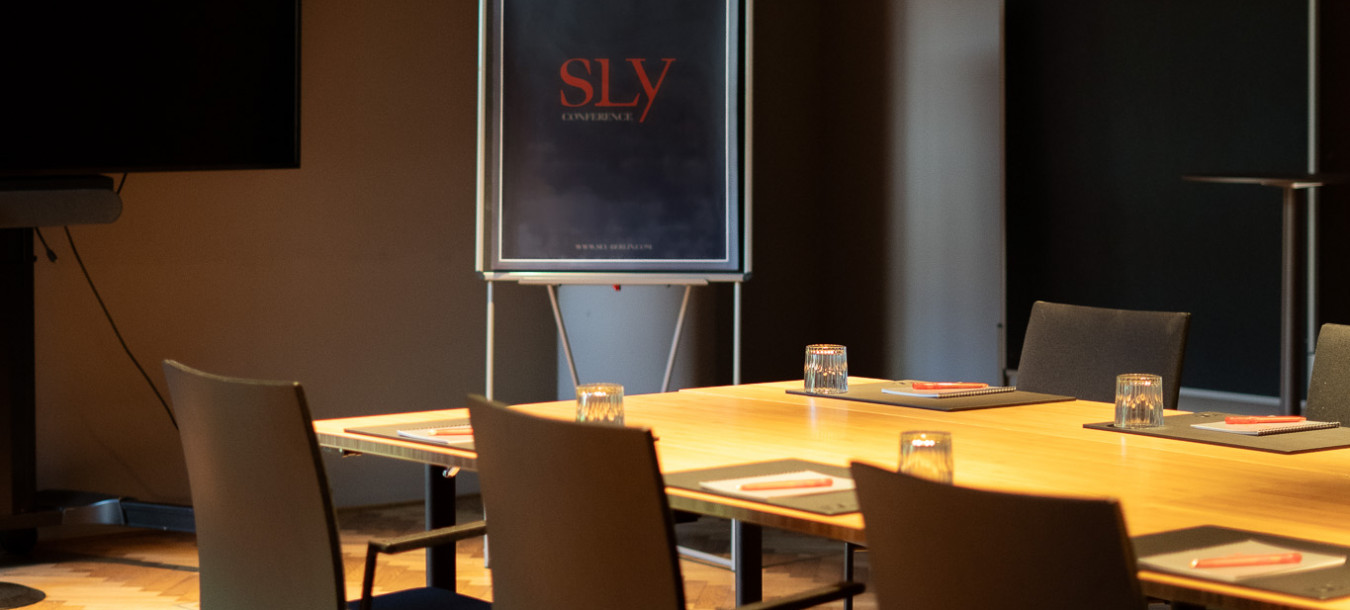 Ein Konferenzraum mit einem rechteckigen Tisch, Stühlen, Gläsern und einem Flipchart, auf dem das Wort SLY in roter Schrift steht.