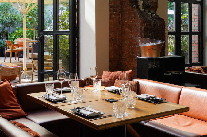 A cozy four-person table in the east Restaurant, with glasses, plates, and napkins, sits on brown leather seats. Sunlight streams in through large windows, and an outdoor terrace is visible in the background.