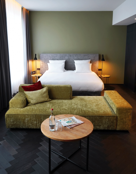 Modernes Hotelzimmer mit großem Bett, grünem Sofa, rundem Holztisch, Wasserflasche und Zeitschriften, sanft beleuchtet durch Nachttischlampen.