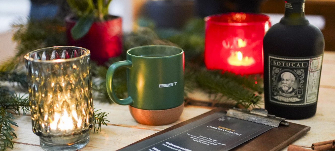 A festive table with candles, a green mug, a bottle, a menu, and greenery, with blurred people in the background.