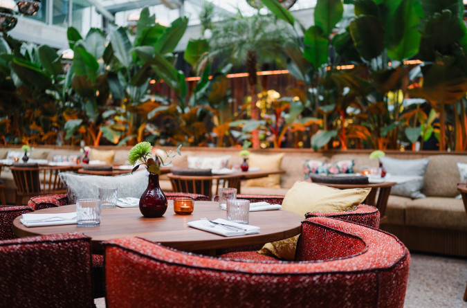 The stylish restaurant interior of the sly Hotel features round wooden tables, red upholstered chairs, and sofas. The tables are set with glasses, cutlery, and flower vases. Lush green plants fill the background.