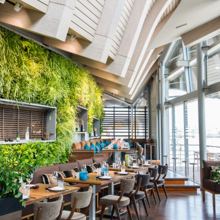Restaurant coast by east mit modernem Interieur mit üppiger vertikaler Gartenmauer und großen Fenstern, die viel Tageslicht hereinlassen. Der Essbereich ist mit Holztischen, gepolsterten Stühlen und dekorativen Kissen ausgestattet und schafft so eine g