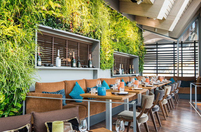 A modern restaurant, coast by east, features a lush green wall unit, brown booths with colorful blue cushions, wooden tables with glassware, and natural light coming in through large windows and skylights.