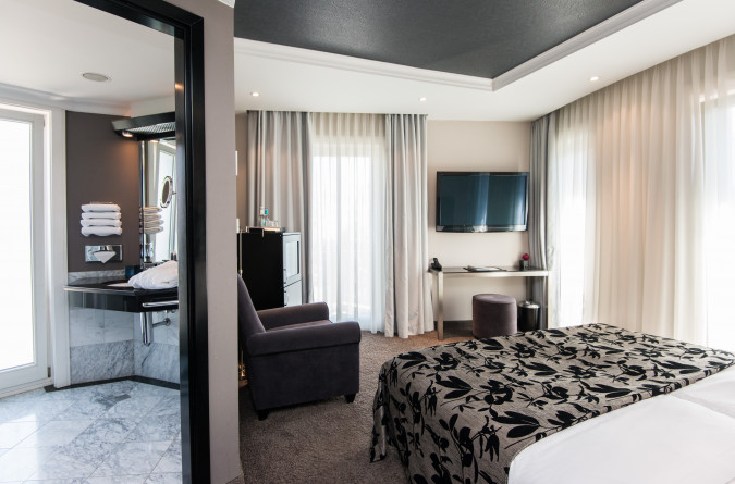 Modernes Zimmer des Hotels New Berlin mit einem Doppelbett mit floralem Muster. An der Wand gegenüber dem Bett hängt ein Fernseher. Es gibt einen Sessel, und das Badezimmer ist teilweise einsehbar und verfügt über Handtücher und eine Waschtischplatte