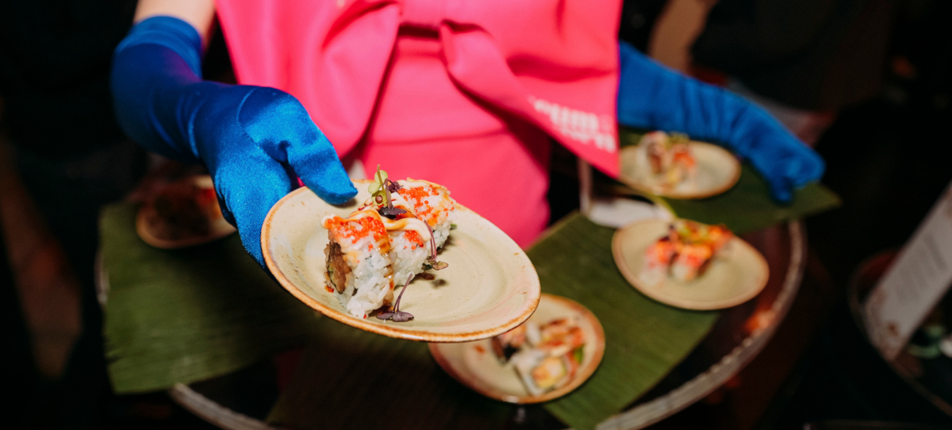 Eine Person mit blauen Handschuhen hält einen Teller mit verschiedenen Sushi-Sorten hoch und präsentiert dessen leuchtende Farben und vielfältige Zutaten.