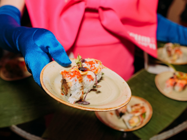 Eine Person mit blauen Handschuhen hält einen Teller mit verschiedenen Sushi-Sorten hoch und präsentiert dessen leuchtende Farben und vielfältige Zutaten.