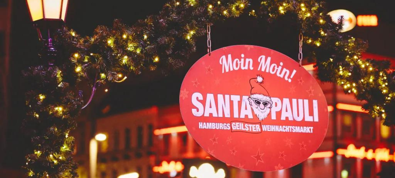 A festive sign reading Santa Pauli hangs on a garland arch, decorated with lights at a Christmas market in Hamburg.