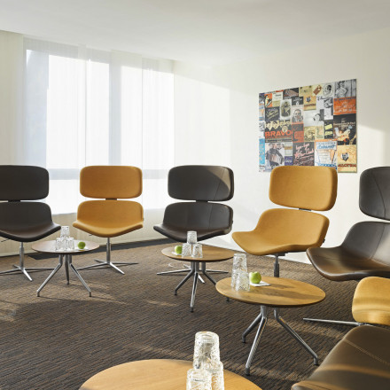 Moderner Konferenzraum des east Hotels mit bunten Polsterstühlen und runden Holztischen. Auf jedem Tisch steht ein Glas Wasser und ein grüner Apfel. An der Wand hängt eine Collage aus Vintage-Postern, und große Fenster lassen Tageslicht herein.