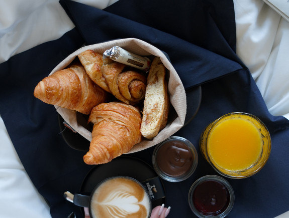 Eine Hand hält eine Kaffeetasse neben Croissants, Brot, Orangensaft und Brotaufstrichen auf einer schwarzen Serviette, alles auf weißer Bettwäsche.