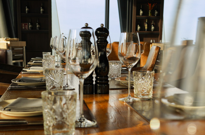A close-up of an elegant restaurant table at clouds Restaurant, set with crystal glasses, wine glasses, plates, silver cutlery, and salt and pepper mills, while sunlight streams through large windows in the background.