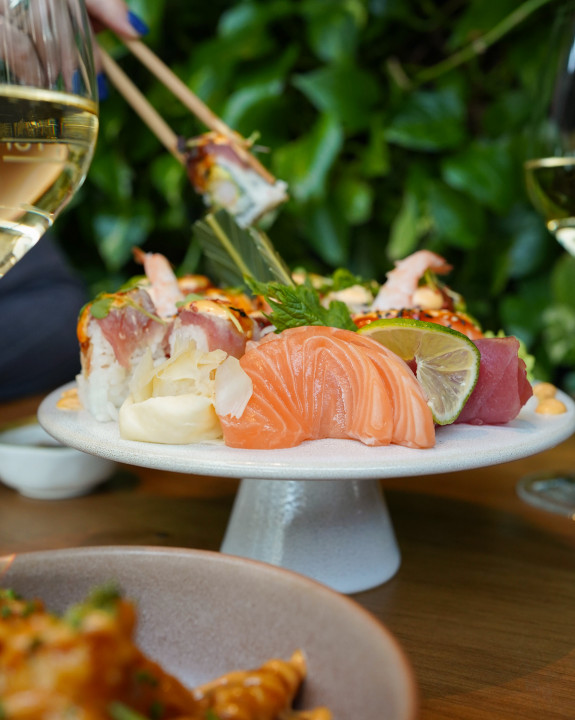Ein Teller mit verschiedenen Sushi- und Sashimisorten mit Garnituren, umgeben von Gläsern mit Weißwein und einem grünen Blatthintergrund.