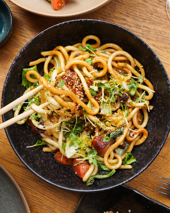 Eine Schüssel mit gebratenen Udon-Nudeln mit Gemüse und Sesamsamen, die mit Stäbchen auf einem Holztisch aufgenommen werden.
