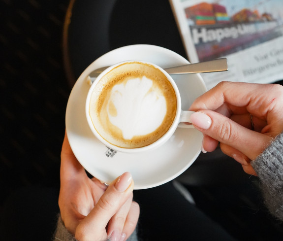 Eine Person hält eine Tasse Cappuccino mit Latte Art neben einer Untertasse, einem Löffel und einer Zeitung auf einem schwarzen Tisch.