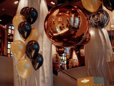 Schwarze und goldene Luftballons und große goldene Dekorationen in einem modernen, stilvollen Restaurant mit weißen Vorhängen und Buntglasfenstern.