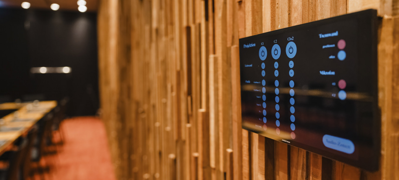 Ein digitales Bedienfeld an einer Wand mit Holzstruktur in einem modernen, schwach beleuchteten Konferenzraum.