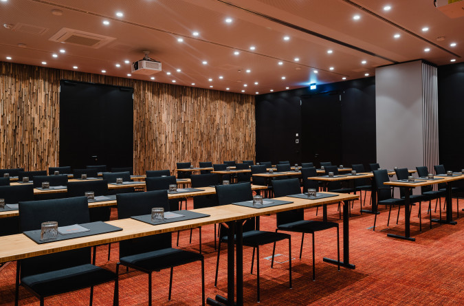 Ein moderner Konferenzraum im sly Hotel mit Tisch- und Stuhlreihen, jeweils mit Tischset, Glas und Stift. Der Raum verfügt über eine warme Holzvertäfelung an einer Wand, Einbauleuchten und einen roten Teppichboden.