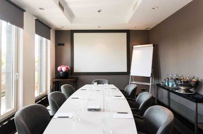 Ein moderner Konferenzraum im Hotel New Berlin mit einem langen weißen Tisch, acht Stühlen, einem leeren Whiteboard, einem Flipchart, Notizblöcken, Gläsern, Getränkeflaschen und einer Vase mit rosa Blumen in der Nähe großer Fenster.