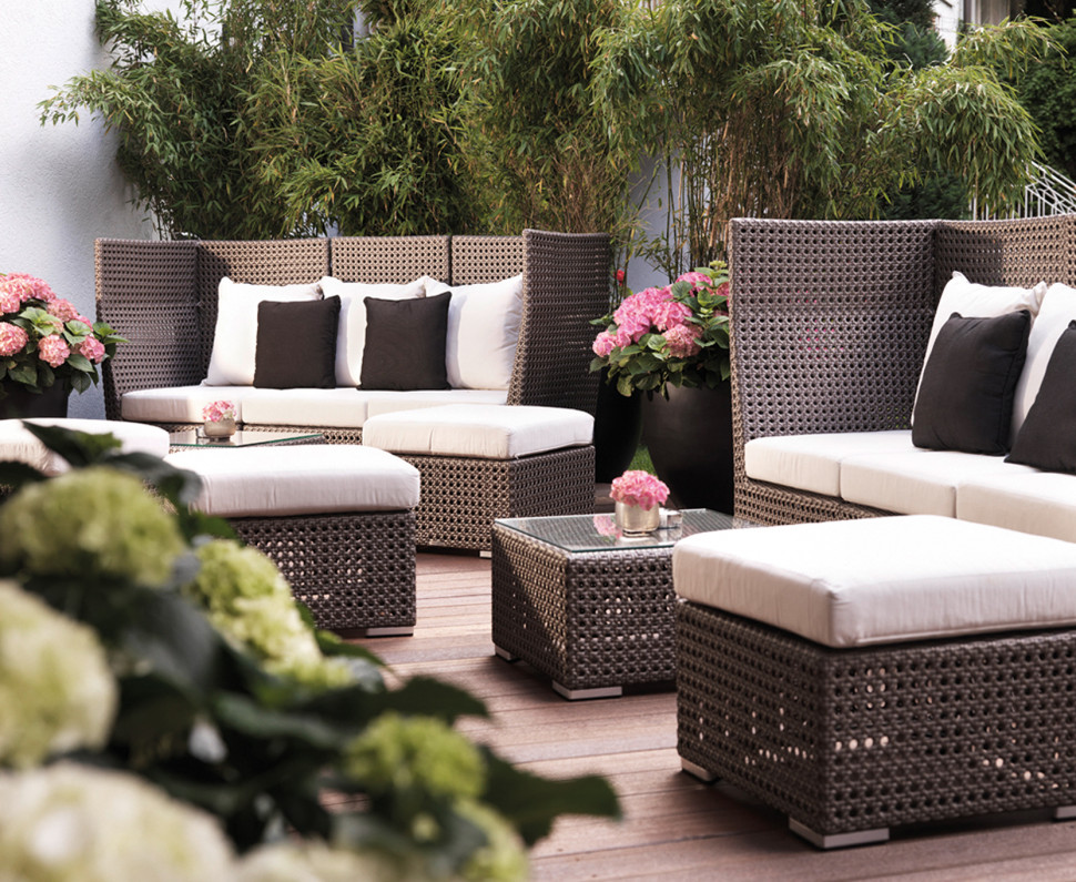 Modern outdoor patio with wicker furniture, white cushions, black and white pillows, potted flowers, and green plants in the background.