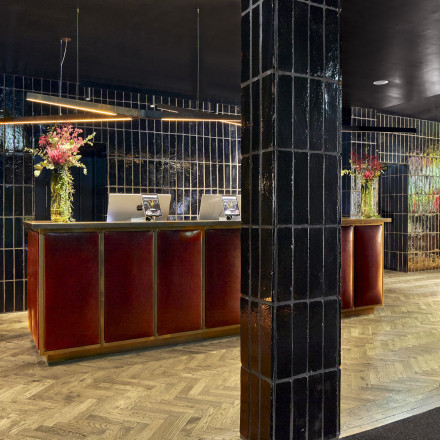Der moderne Rezeptionsbereich des sly Hotels besticht durch einen langen roten Schreibtisch mit Laptops und Blumenarrangements. Wände und eine Säule sind mit glänzenden dunklen Fliesen verkleidet, der Boden ist mit einem Fischgrätenmuster versehen. De
