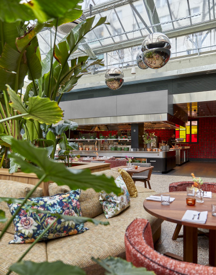 Ein modernes Restaurant mit tropischen Pflanzen und farbenfrohem Dekor. Runde Tische und gepolsterte Sitzgelegenheiten sind rund um die offene Küche angeordnet. Große silberne Kugelleuchten hängen von der Glasdecke. Bunte Kissen sorgen für eine gemüt
