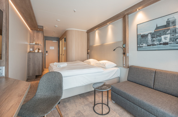 Hotelzimmer des Park Inn Wismar mit Doppelbett, grauem Polstersofa, kleinem runden Tisch und grauem Stuhl. Minimalistisches Dekor mit Holzakzenten und einem Stadtbild an der Wand. Ein kleiner Kühlschrank und eine Kaffeestation befinden sich in der Nähe 