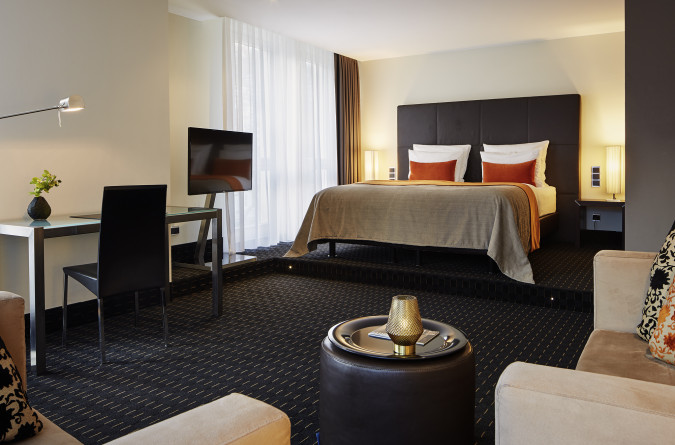 Hotelzimmer im Boston Hotel mit einem großen Bett mit braun-orangefarbener Bettwäsche, einem Schreibtisch mit Lampe und Stuhl, einem Sofa und einem Fernseher. Im Vordergrund steht ein runder Tisch mit einem Deko-Objekt. Ein großes Fenster mit transpare