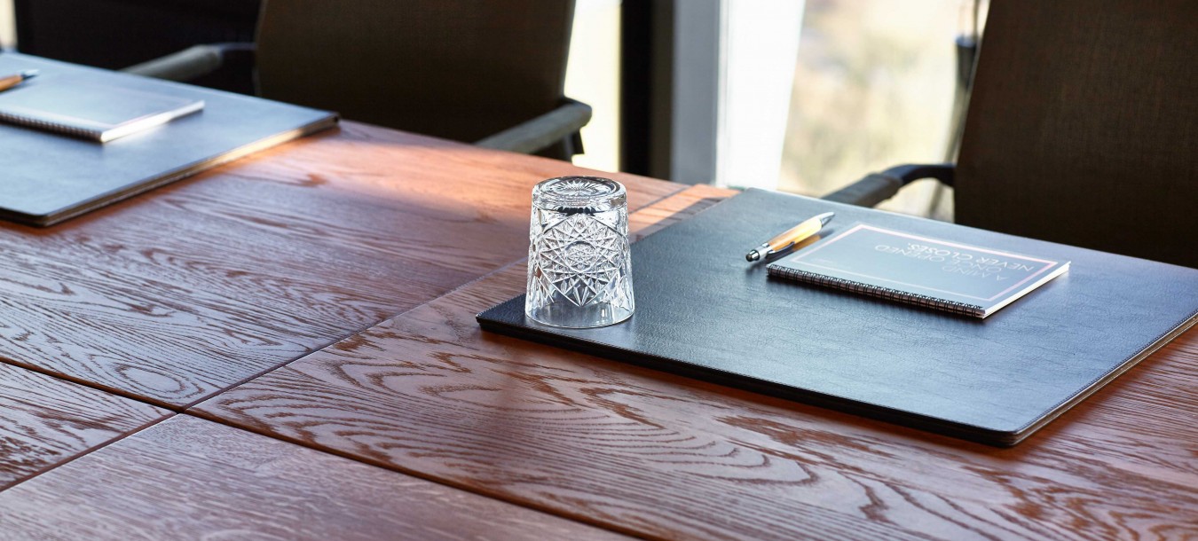Nahaufnahme eines Konferenztisches mit schwarzen Tischsets, Notizbüchern, Stiften und einem umgedrehten Glas auf der Holzplatte. Um den Tisch herum stehen leere Stühle.
