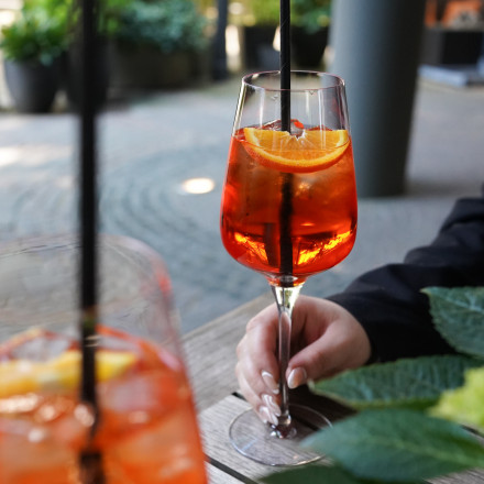 Eine weibliche Person hält ein Glas orangefarbenen Cocktail mit Eis und Orangenscheiben auf einem Holztisch auf einer Terrasse mit grünen Pflanzen im Vordergrund. Ein weiteres ähnliches Getränk ist im unscharf im Vordergrund sichtbar.