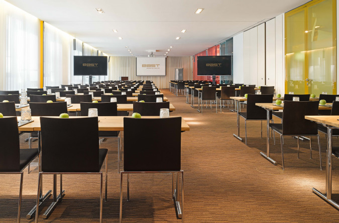 Ein geräumiger, moderner Konferenzraum im east Hotel mit Tischreihen und schwarzen Stühlen. Auf jedem Tisch steht ein Glas und ein grüner Apfel. Im vorderen Teil des Raumes sind zwei große Bildschirme und eine Projektionsleinwand zu sehen.
