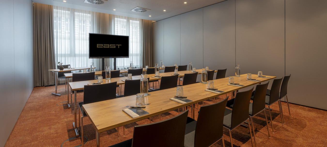 Konferenzraum mit Tisch- und Stuhlreihen, einem Fernsehbildschirm mit Ost-Ausrichtung und natürlichem Licht durch große Fenster.