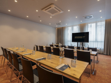 Ein moderner Konferenzraum mit Tisch- und Stuhlreihen, einem Fernsehbildschirm an der Vorderseite und großen Fenstern mit Vorhängen.