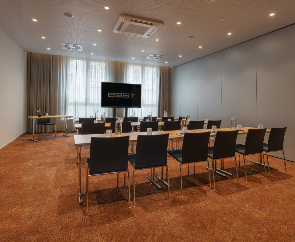 Ein moderner Konferenzraum mit Stuhlreihen, Tischen, einer großen Leinwand und natürlichem Licht durch hohe Fenster.