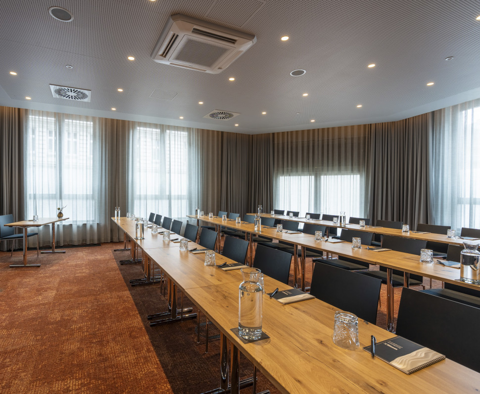 Modern conference room with long tables, chairs, water pitchers, glasses, notepads, and a large screen, with daylight streaming through windows.