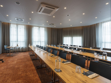 Modern conference room with long tables, black chairs, a large screen, and natural light from tall windows with beige curtains.