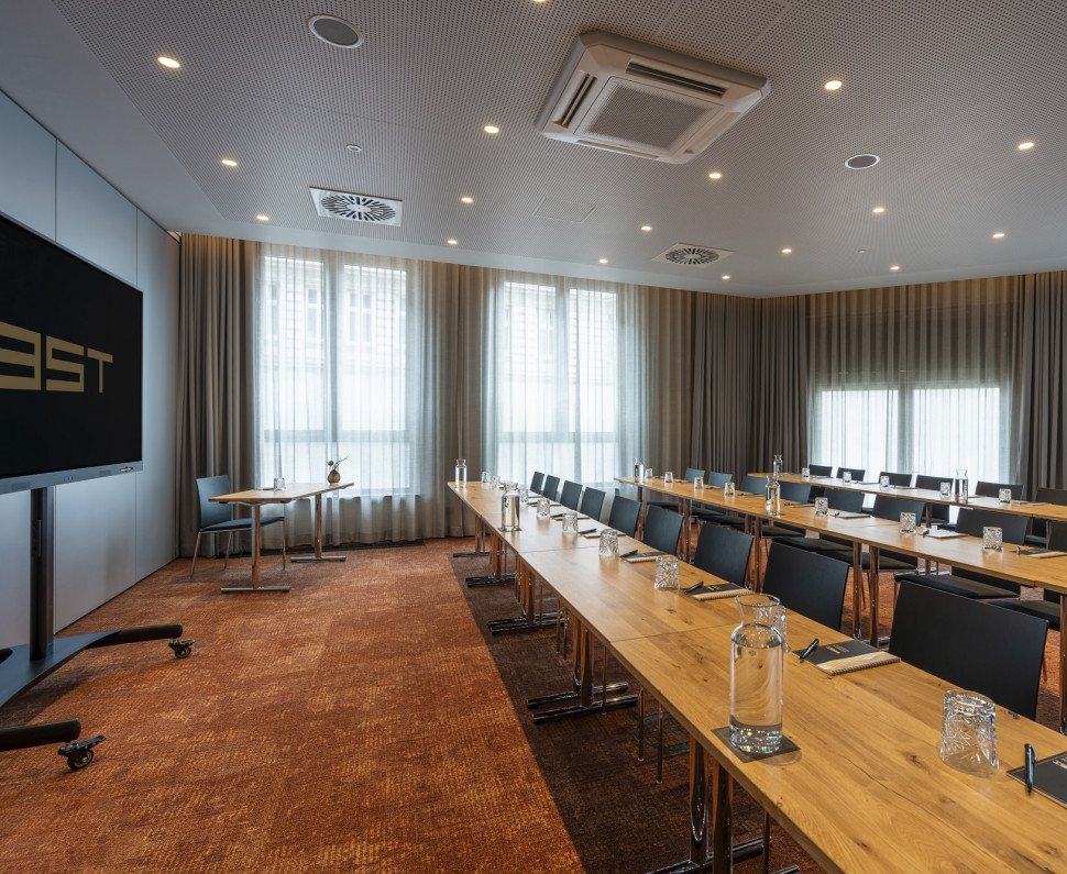 Ein moderner Konferenzraum mit langen Tischen, Stühlen, Notizblöcken, Gläsern und einem großen Bildschirm, bereit für ein Meeting oder Seminar.