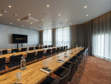 Moderner Konferenzraum mit Holztischen, schwarzen Stühlen, Wasserkrügen, Gläsern, Notizblöcken und einem großen Bildschirm davor.