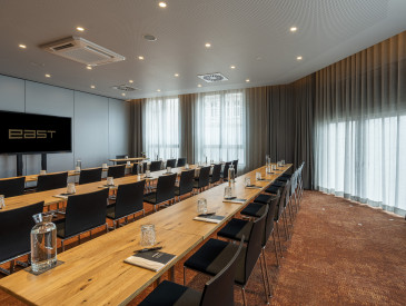 Modern conference room with long wooden tables, black chairs, notepads, glasses, and a TV screen displaying EAST, with large windows and curtains.