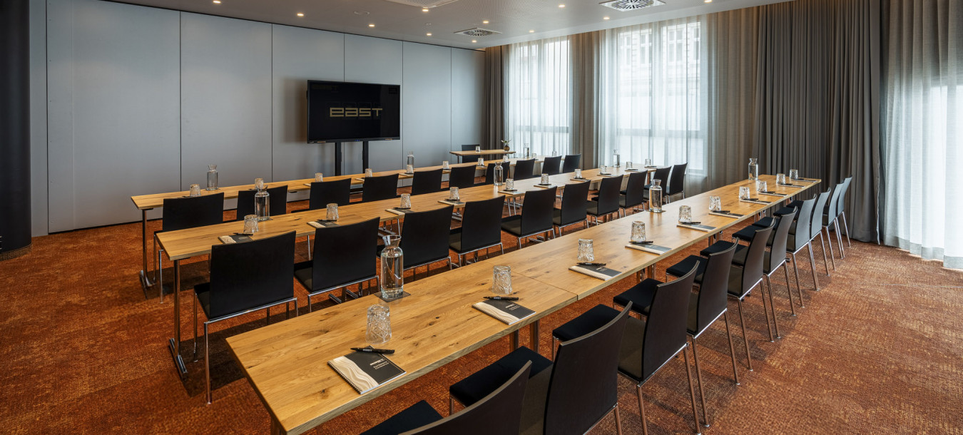 Moderner Konferenzraum mit Tisch- und Stuhlreihen, Notizblöcken und Wasserflaschen vor einem großen Bildschirm und Fenstern mit Vorhängen.