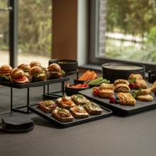 Ein gedeckter Tisch mit Mini-Sandwiches, Toast, Keksen, Kuchen, frischem Gemüse und schwarzen Schüsseln in der Nähe großer Fenster.