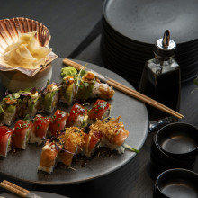 Ein Teller mit verschiedenen Sushi-Rollen mit Garnitur, eine Schale mit eingelegtem Ingwer, Sojasauce und schwarze Teller, gestapelt auf einem dunklen Tisch.