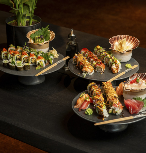 Drei Teller mit verschiedenen Sushi-Rollen und Sashimi auf einem dunklen Tisch, mit Essstäbchen und einer kleinen Topfpflanze im Hintergrund.