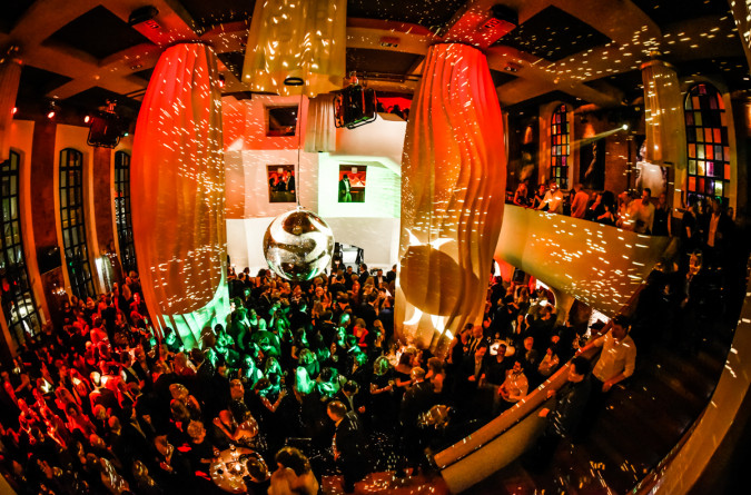 Eine gut besuchte, lebhafte Party im east Hotel in einem großen, modernen Veranstaltungsort mit farbenfroher Beleuchtung, einer Discokugel, die das Licht reflektiert, und Menschen, die im Erdgeschoss und auf den Balkonen tanzen und sich unterhalten.